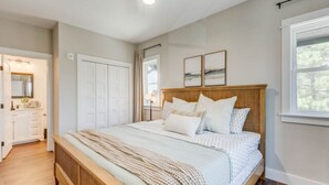 A bedroom with a wooden bed, white bedding, a large window, and a door leading to a bathroom. - The Downtown Delight Family Suite (Manteo)