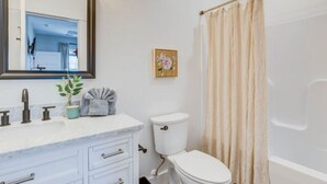 A bathroom with a white toilet, a sink with a marble countertop, a mirror, and a shower curtain. - The Downtown Delight Family Suite (Manteo)