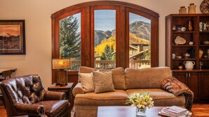 A cozy living room with a leather armchair, a brown sofa with cushions, a wooden coffee table, and a bookshelf. A large window offers a view of mountains and trees. - Beautiful Sun Valley Lodge II Penthouse, sleeps 8 with amazing views of Baldy (Sun Valley)