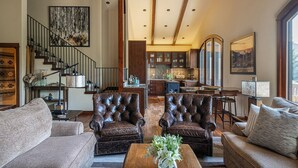 A spacious living area with leather armchairs, a sofa, a coffee table with flowers, a staircase, a kitchen, and large windows. - Beautiful Sun Valley Lodge II Penthouse, sleeps 8 with amazing views of Baldy (Sun Valley)