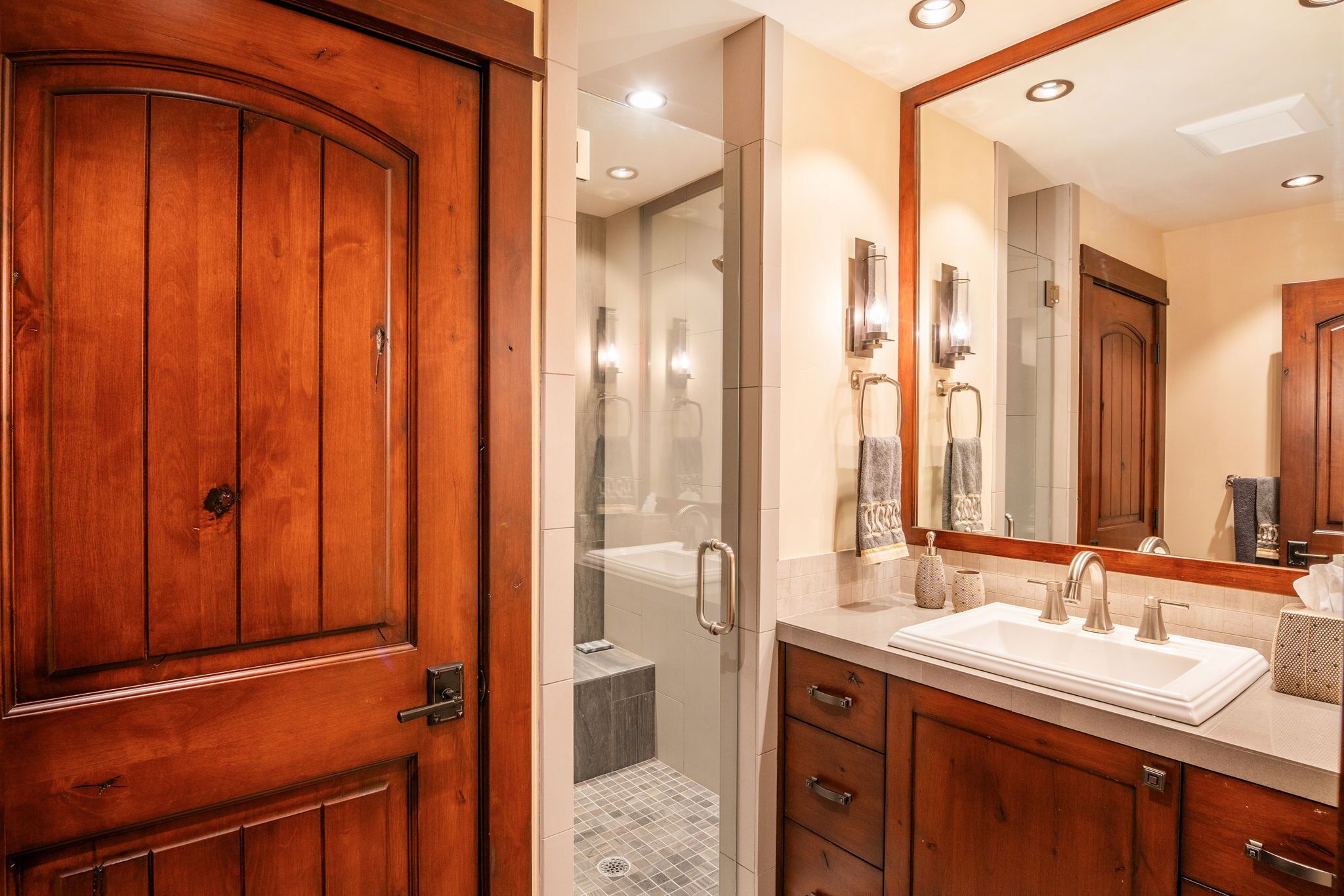 A bathroom with a wooden door, a large mirror, a sink, and a shower area.
