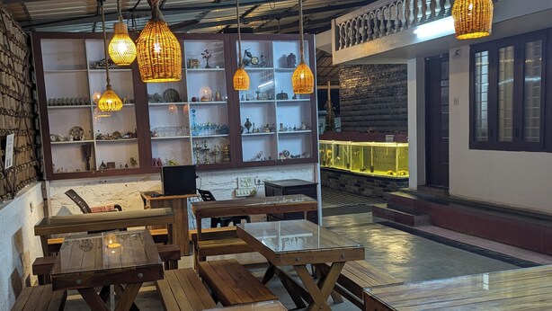 A cozy restaurant with wooden tables and benches, hanging wicker lamps, and shelves displaying various items. - NatureVilla Kochi (Aluva)