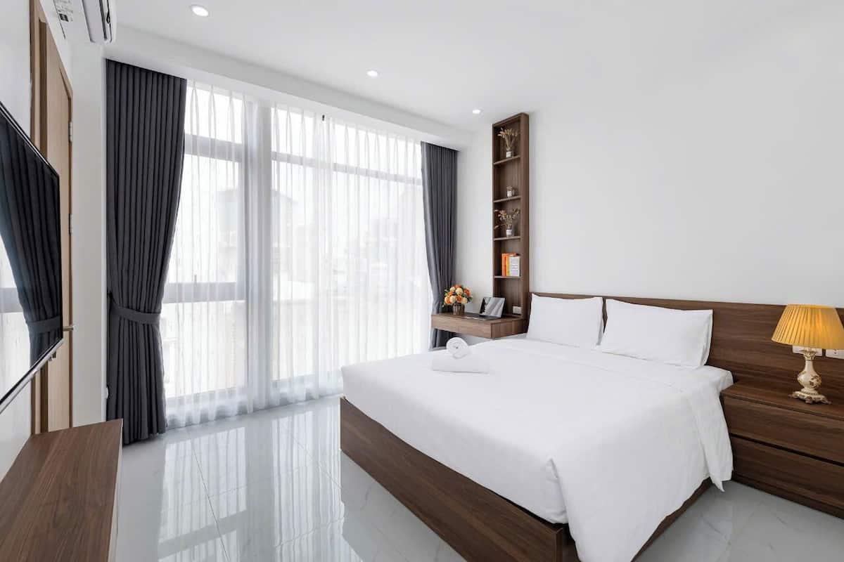 A modern hotel room with a large bed, a bedside table, a nightstand, a TV, a bookshelf, and a window with curtains.