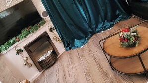 A living room with a teal curtain, a flat-screen TV, a decorative Christmas wreath, and a small wooden side table with a flower arrangement. - Haven Holiday Park 42 Pine Lodge (Pwllheli)