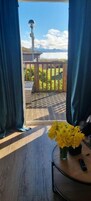 Una habitación con vistas a una terraza y una valla de madera, suelo de madera, una mesa redonda con un florero de flores amarillas y un control remoto.