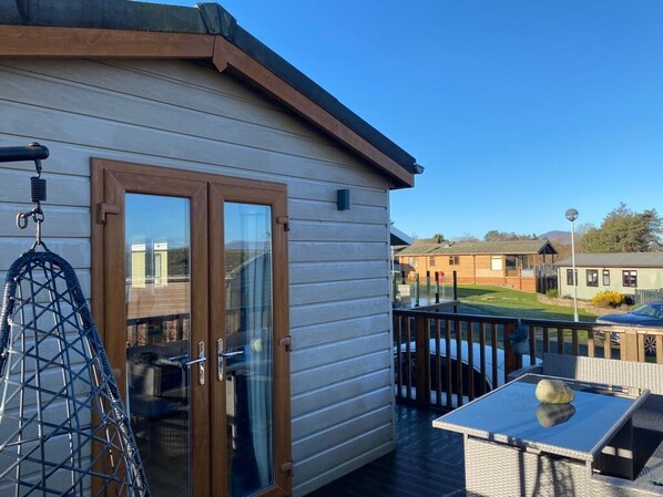 A wooden deck with a glass door leading to a room, a table with a chair, and a hanging chair. - Haven Holiday Park 42 Pine Lodge (Pwllheli)