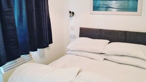 A bedroom with a bed, white pillows, a headboard, a wall-mounted light, a painting of a seascape, and a window with curtains. - Haven Holiday Park 42 Pine Lodge (Pwllheli)