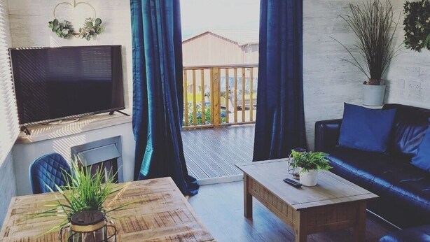 A cozy living room with a wooden table, a sofa, a TV, and a balcony with blue curtains. - Haven Holiday Park 42 Pine Lodge (Pwllheli)