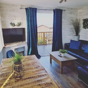Ein gemütliches Wohnzimmer mit Holztisch, einer Couch, einem Fernseher und einem Balkon mit blauen Vorhängen.