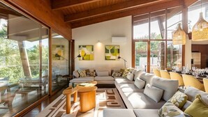 A modern living room with a large sofa, wooden accents, and large windows offering a view of the outdoors. - Ponte de Lima Chalet Pool & Golf (Feitosa)