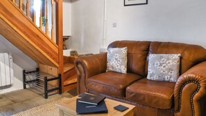 Un salon avec un canapé en cuir marron, deux coussins à motifs floraux, une table basse en bois sur laquelle sont posés des livres, et un escalier avec des rampes en bois.