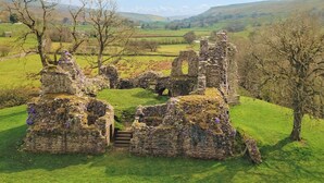 Une ruine historique en pierre, avec un escalier menant à une arche centrale, le tout entouré de champs verdoyants et d’arbres.