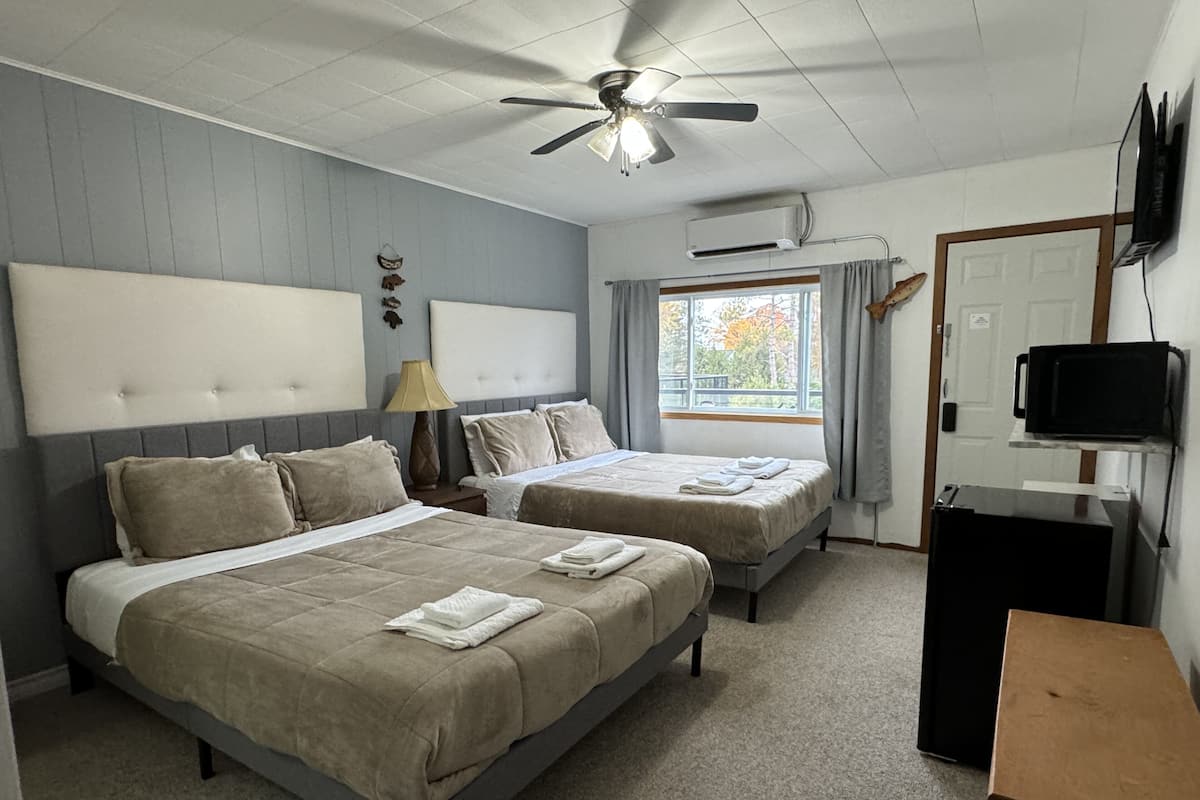 A hotel room with two beds, a ceiling fan, an air conditioning unit, a television, and a window with curtains.