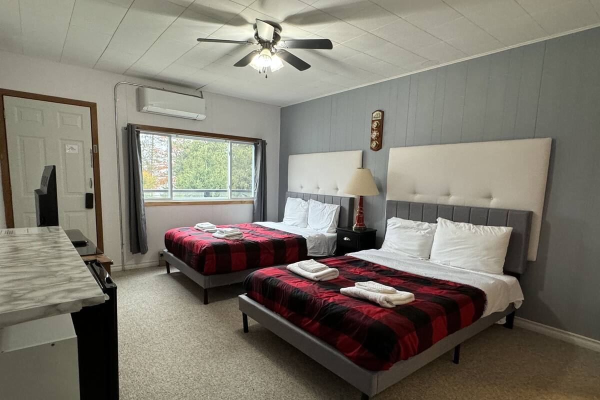 A hotel room with two beds, a ceiling fan, a window with a view of trees, and a clock on the wall.