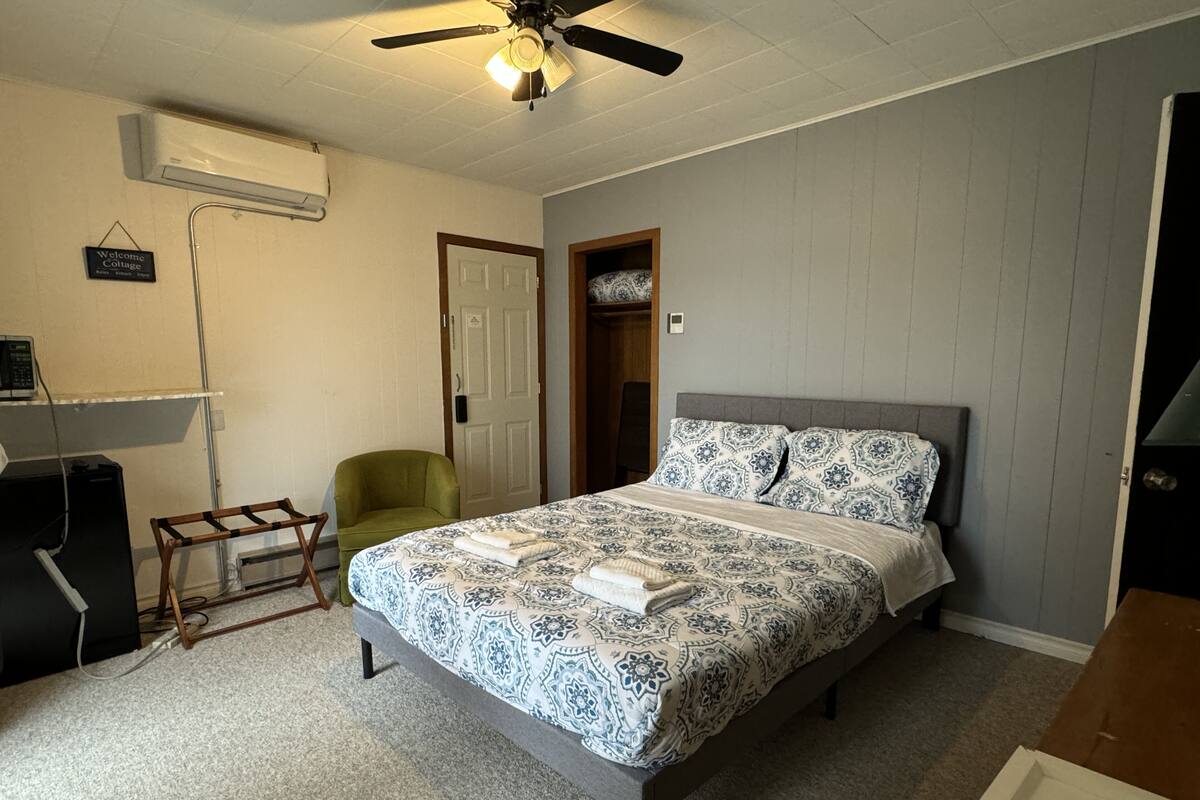 A bedroom with a bed, a ceiling fan, a chair, a door, and a small table.