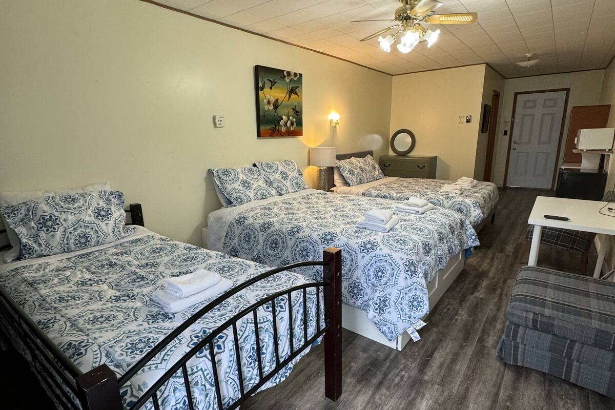 A room with two beds, a small table, a chair, a ceiling fan, and a painting on the wall.