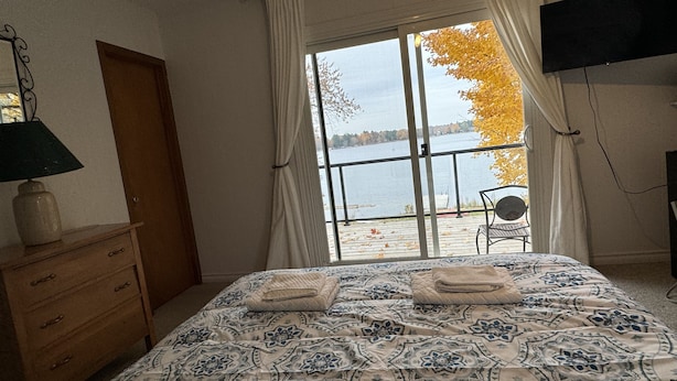 A bedroom with a large bed, a wooden dresser, a lamp, and a view of a lake through a window with curtains. - Lake Sparrow Guest House Inc (Severn Bridge)
