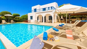 A pool area with lounge chairs, umbrellas, and a building with a white facade and blue accents. - Villa Artemis - Four Bedroom Villa, Sleeps 8 (Peyia)