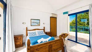 A bedroom with a wooden bed, a painting, a door, and a balcony with a view. - Villa Artemis - Four Bedroom Villa, Sleeps 8 (Peyia)