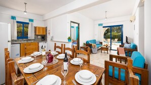 A spacious dining area with a wooden table set for six, a kitchen with wooden cabinets, and a living area with a sofa and chairs. - Villa Artemis - Four Bedroom Villa, Sleeps 8 (Peyia)