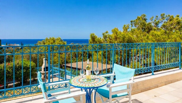 A balcony with a table and chairs, overlooking a scenic view of the sea and greenery.
