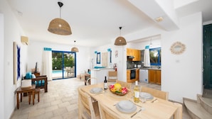 A spacious dining area with a wooden table set for four, a kitchen with wooden cabinets, and a view of a swimming pool through glass doors.