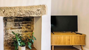 A room with a stone fireplace, a wooden cabinet, and a television on a stand. - Saltaire Mill Cottage (Saltaire, near Haworth)