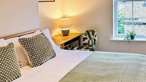 A hotel room with a bed, a nightstand with a lamp, a chair, a window with a view of buildings, and a framed picture on the wall. - Saltaire Mill Cottage (Saltaire, near Haworth)