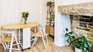 A dining area with a round wooden table, white chairs, a vase with yellow flowers, and a bookshelf with books and decorative items. - Saltaire Mill Cottage (Saltaire, near Haworth)