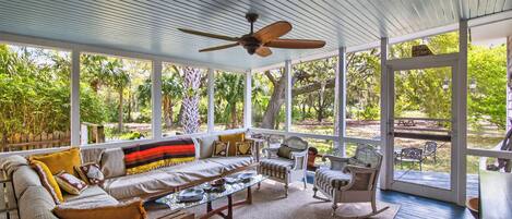 A covered porch with a ceiling fan, a sofa with cushions, wicker chairs, and a view of the garden.