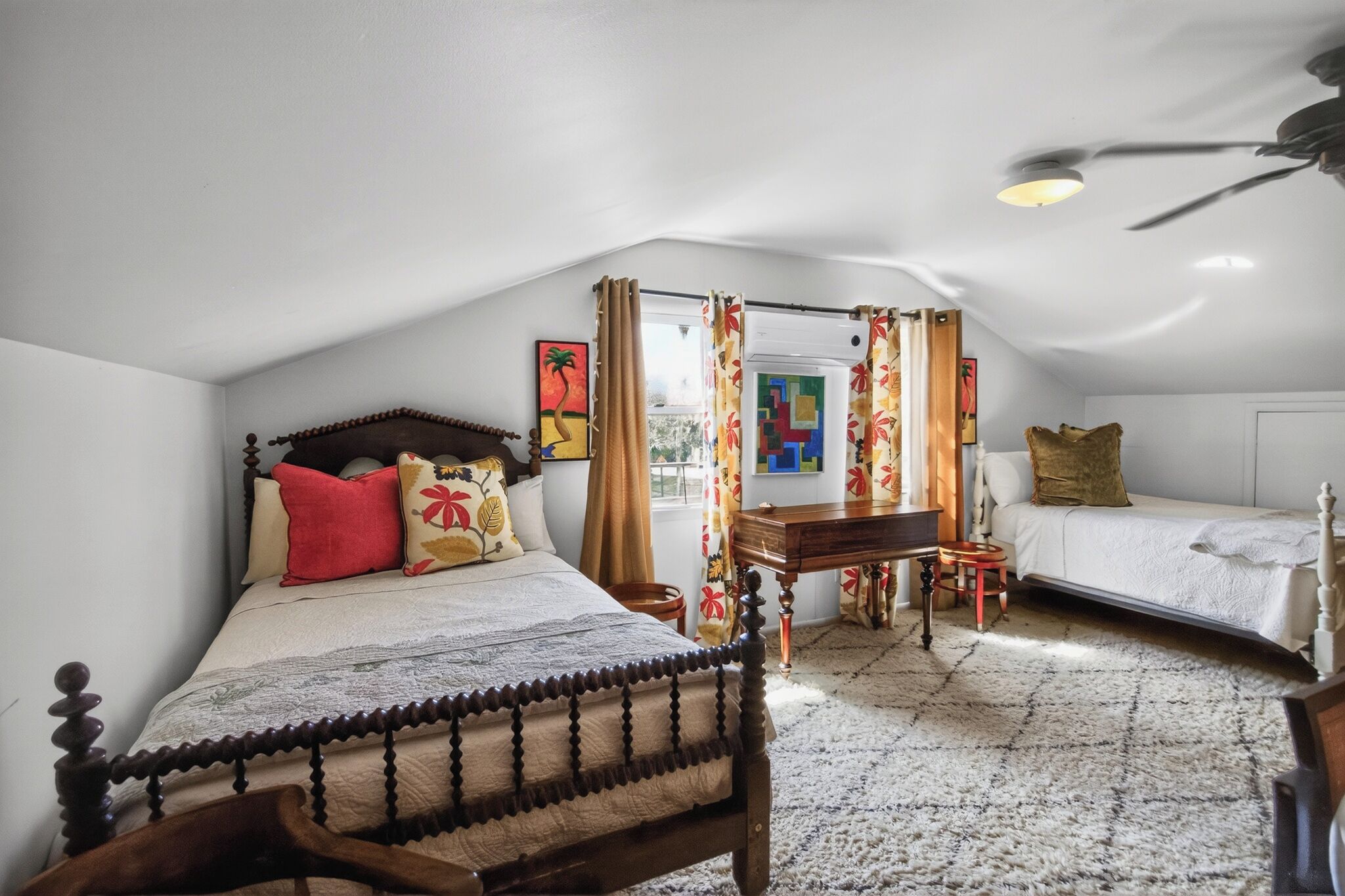 A bedroom with a wooden bed, a desk, and a chair. There is a window with curtains, a ceiling fan, and a painting on the wall.