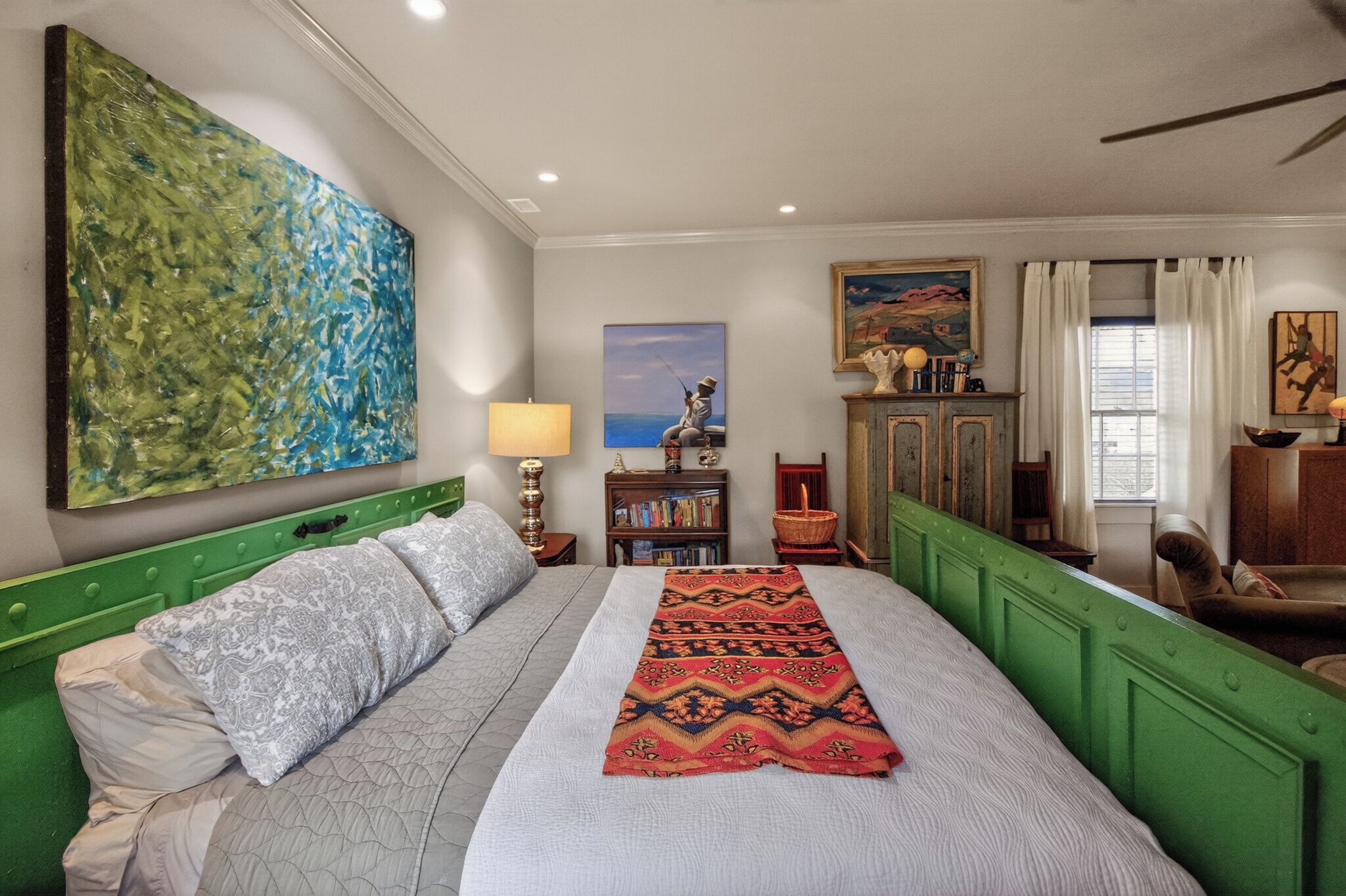 A bedroom with a bed, a green headboard, a painting, a bookshelf, and a window with curtains.