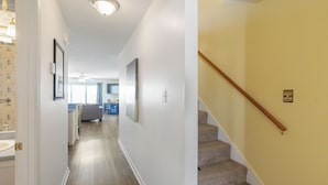 A hallway with a carpeted floor, a staircase with a wooden handrail, and a door leading to a bathroom. - Bart’s Back River Retreat | Spacious 3BR Townhome | Water Views | South End (Tybee Island)