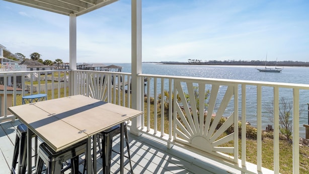 A covered outdoor area with a table and chairs, overlooking a body of water with a boat in the distance. - Bart’s Back River Retreat | Spacious 3BR Townhome | Water Views | South End (Tybee Island)