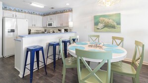 A kitchen with white cabinets, a blue countertop, and a dining area with green chairs and a white table. - Bart’s Back River Retreat | Spacious 3BR Townhome | Water Views | South End (Tybee Island)