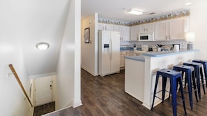 A kitchen with white cabinets, a refrigerator, a microwave, and a bar area with stools. - Bart’s Back River Retreat | Spacious 3BR Townhome | Water Views | South End (Tybee Island)