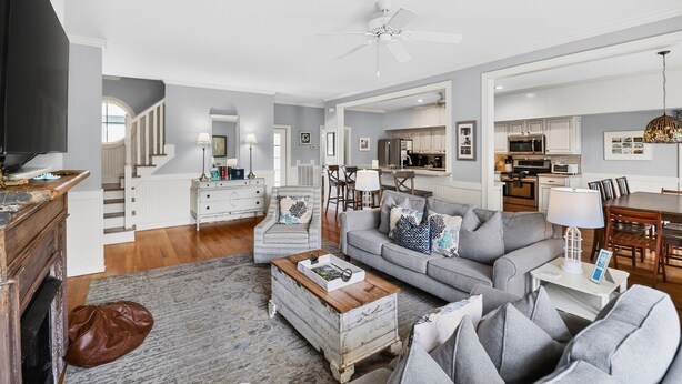 A spacious living room with a grey sofa, coffee table, and a fireplace. A staircase leads to a kitchen area with white cabinets and a dining table. - Large House w Pool Table, close Restaurants + Beach Access less than 1000 FT (Tybee Island)