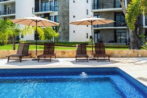 Piscina con sillas de descanso y sombrillas, rodeada de un jardín ajardinado y un edificio moderno al fondo.