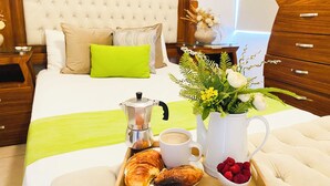 Ein ordentlich bezogenes Bett mit einem grünen Kopfkissen, ein Nachttisch mit einer Kaffeemaschine, eine Tasse Kaffee, Gebäck und eine Vase mit Blumen.