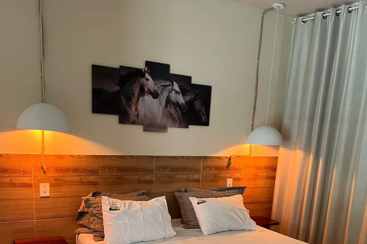 A hotel room with a bed, two bedside lamps, a headboard with artwork, and a window with curtains.