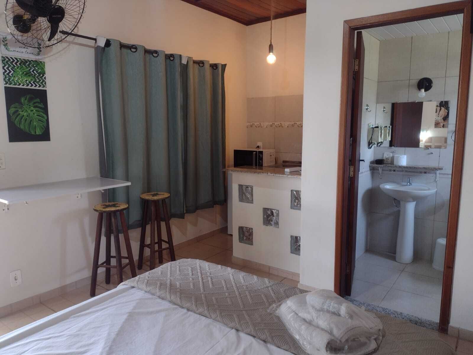 Une chambre comprenant un lit, un petit bureau, deux tabourets, un coin cuisine et une salle de bain avec un lavabo et un miroir.