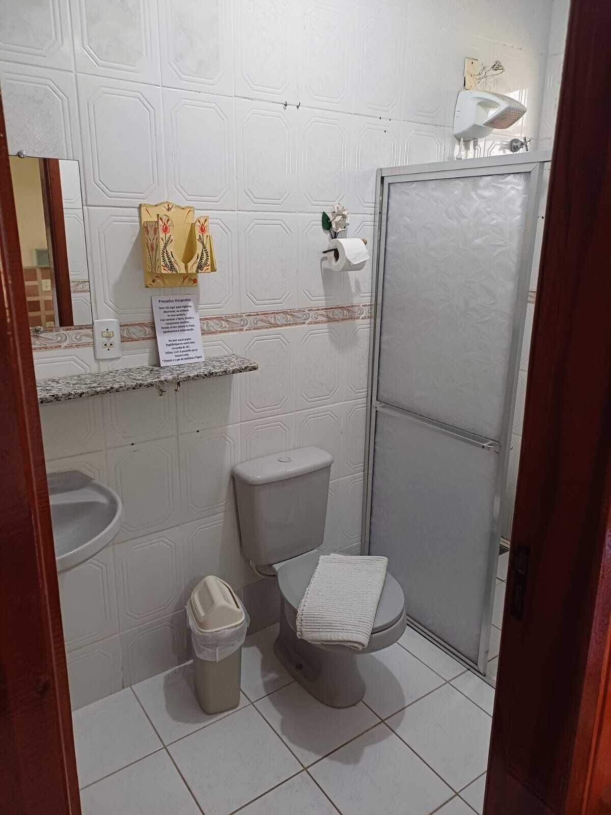 Un baño pequeño con inodoro, lavamanos y espejo. Se ve un dispensador de toallas de papel y un cubo de basura.