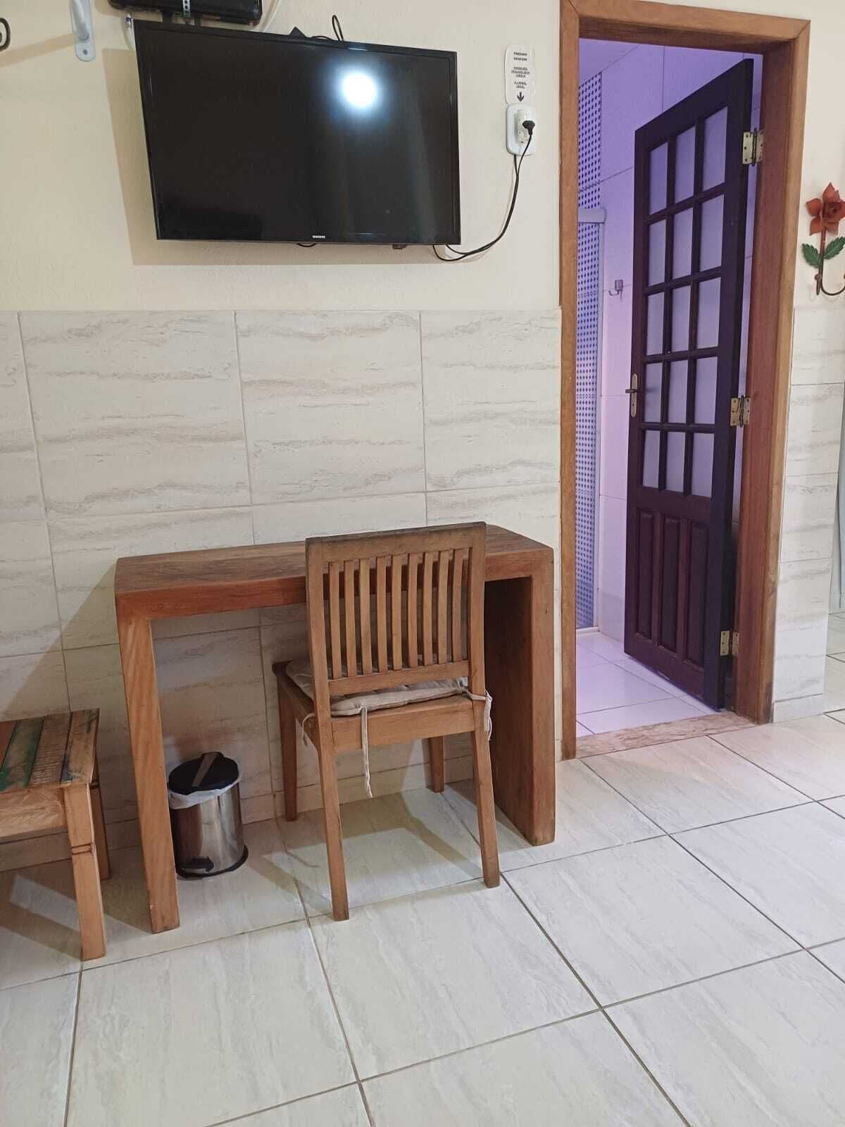 A room with a wooden desk, a chair, a small table, a trash bin, and a television mounted on the wall.