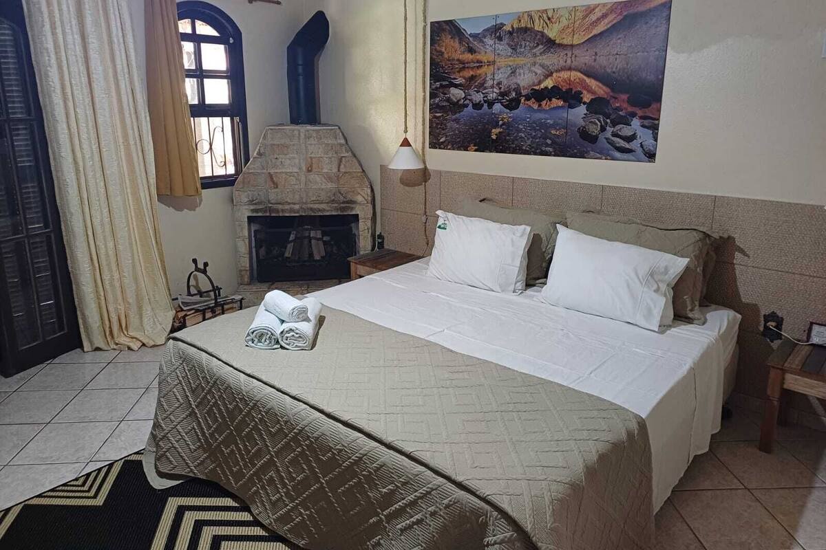 A bedroom with a stone fireplace, a bed with white linens, a painting of a mountainous landscape, and a window with curtains.