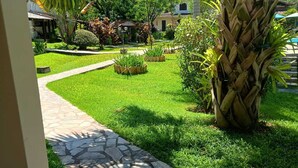 Un sentier de jardin mène à un bâtiment doté d’un toit en tuiles, entouré d’une végétation luxuriante et d’un palmier.