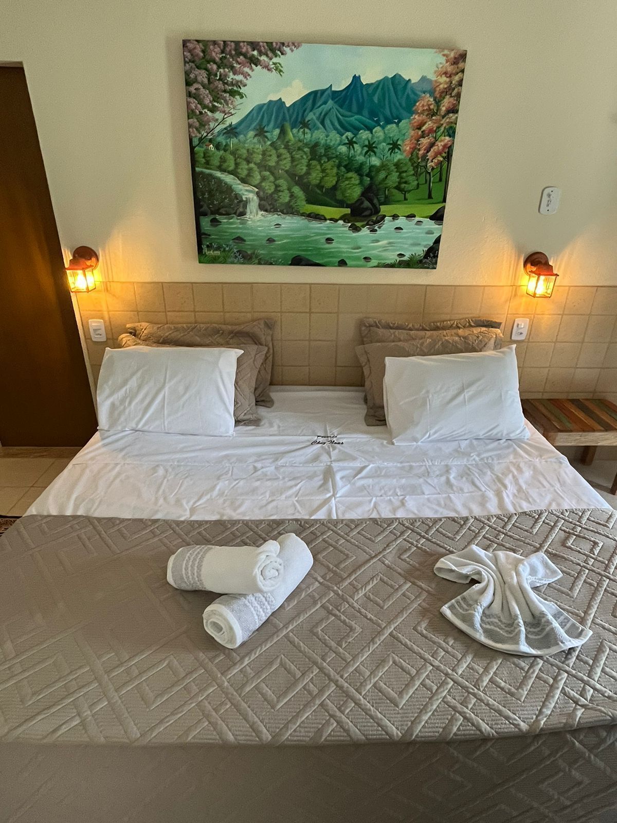 Habitación de hotel con cama, dos almohadas, un cuadro de un paisaje y dos lámparas de pared.