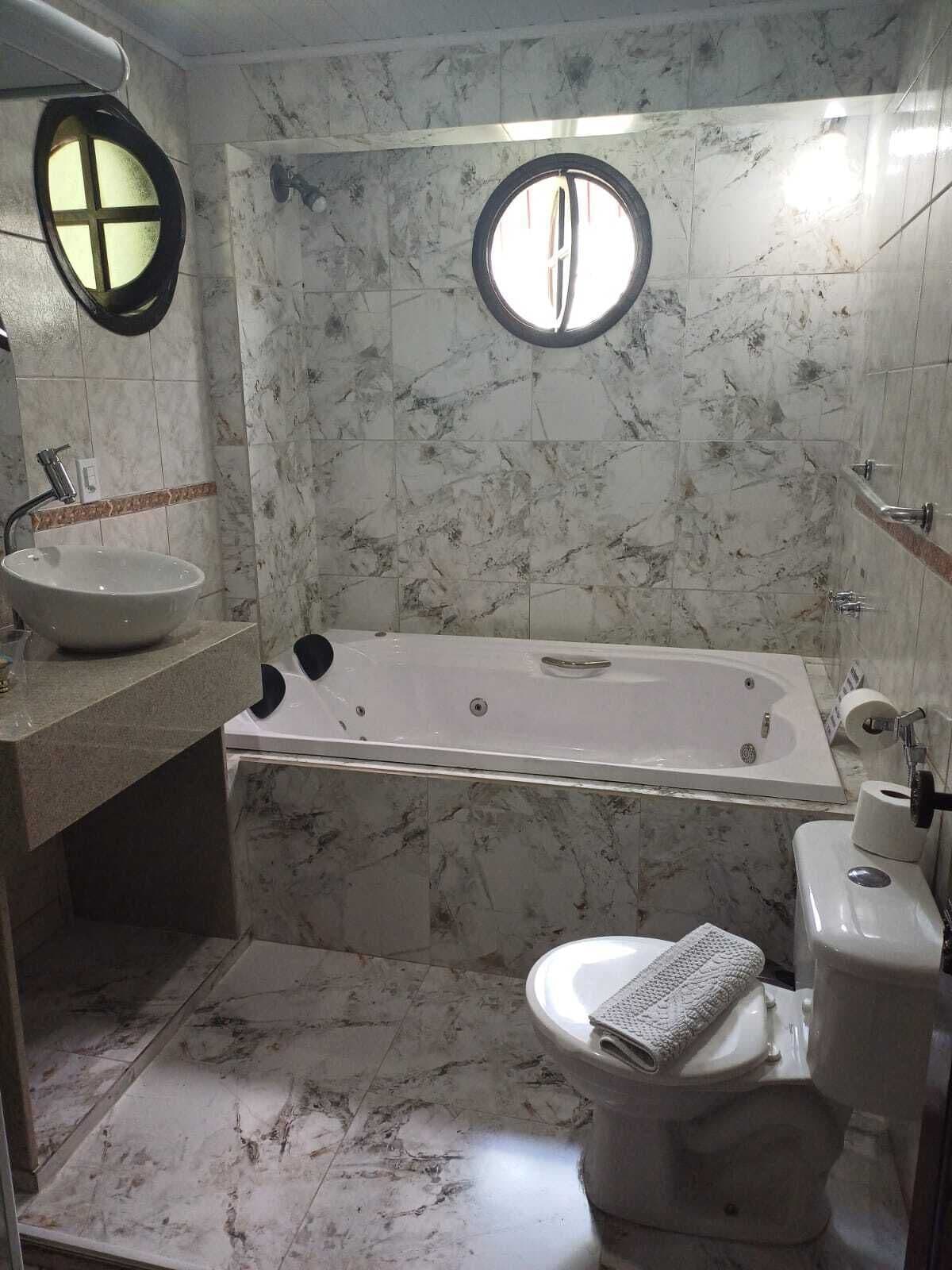 Un baño con bañera, inodoro, lavabo y dos ventanas redondas.