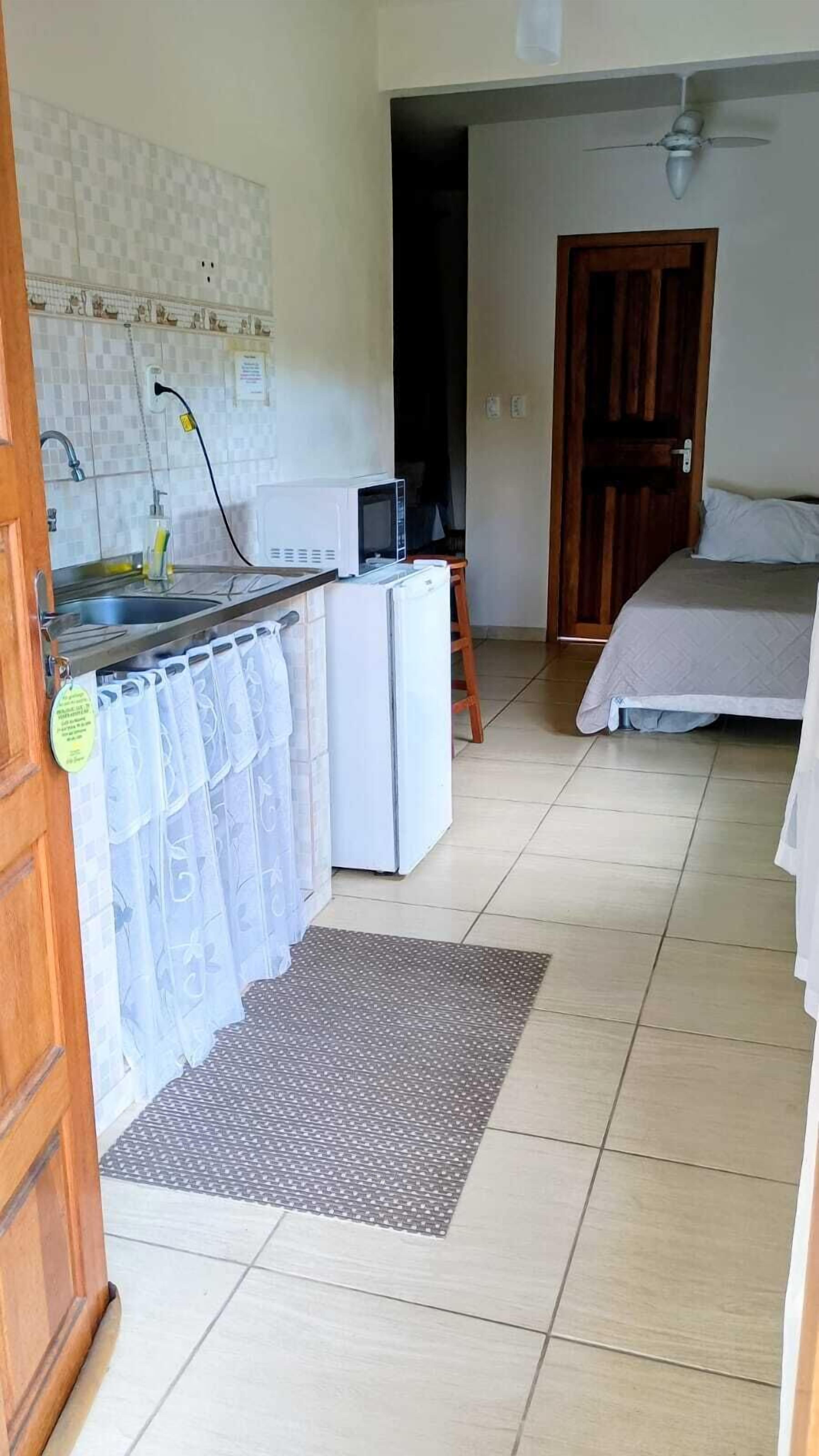 A compact kitchen with a microwave, refrigerator, and sink. There is a small wooden stool and a bed with a white bedspread in the adjacent room.