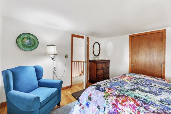 A bedroom with a blue armchair, a bed with a colorful quilt, a wooden dresser, a mirror, and a green plate on the wall.