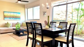 Un comedor con una mesa negra, cuatro sillas y una araña. Se observa un sofá con cojines al fondo.